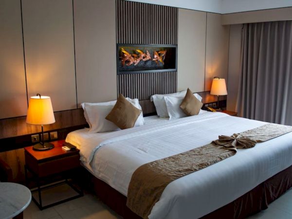 This image shows a neatly arranged hotel room with a king-size bed, wall-mounted lamps on nightstands, and a small table by a window with curtains.