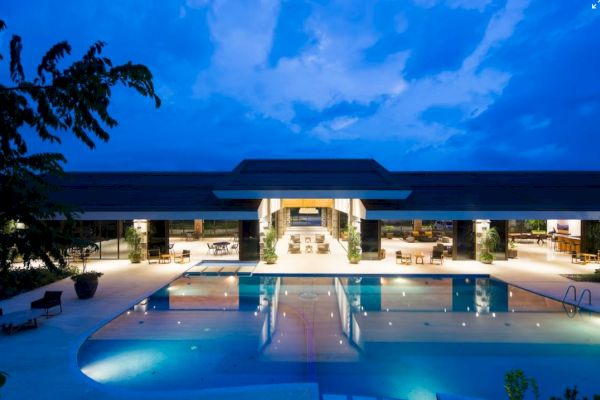 A luxurious swimming pool with a modern, well-lit building in the background, taken during twilight under a blue sky with scattered clouds.