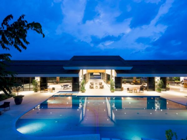 A luxurious swimming pool with a modern, well-lit building in the background, taken during twilight under a blue sky with scattered clouds.