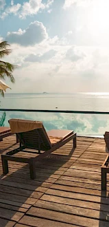 A serene poolside scene with lounge chairs, palm trees, parasols, and a calm body of water reflecting the sky at sunset.
