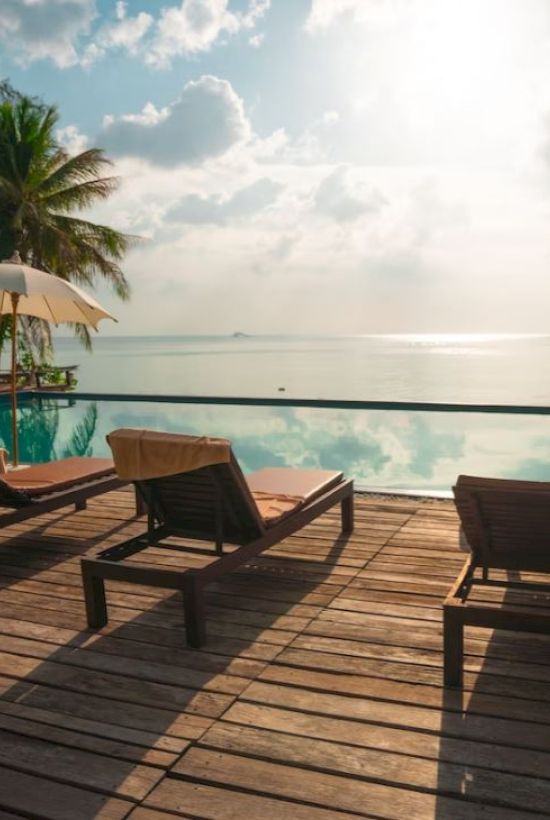 A serene setup with lounge chairs and umbrellas by an infinity pool overlooking a calm sea, framed by palm trees under a partly cloudy sky.