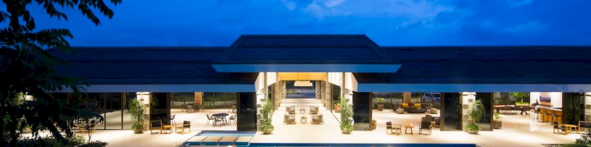 A modern building with illuminated interiors overlooks a spacious, lit-up swimming pool under an evening sky, surrounded by trees and seating areas.