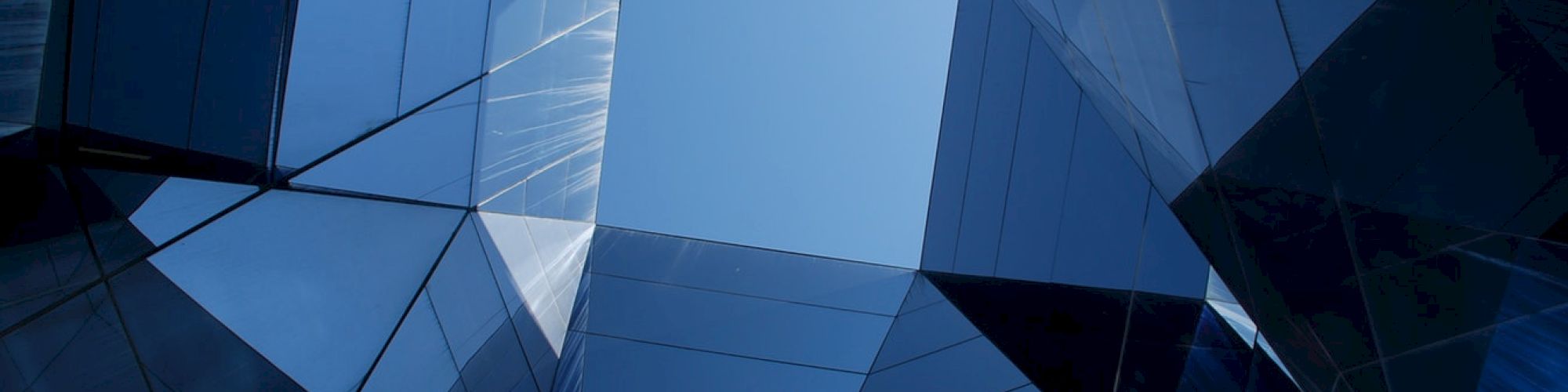 The image shows angular, reflective glass surfaces from a modern building structure creating a geometric pattern against the open blue sky.