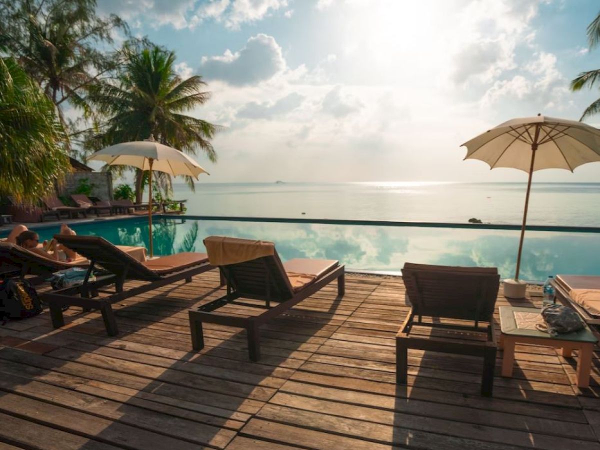 A serene poolside scene with lounge chairs and umbrellas on a wooden deck overlooking an infinity pool and a tranquil sea with a bright sky.