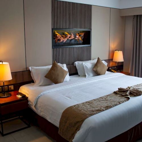 A hotel room with a large bed, bedside tables with lamps, a wall decoration, and a curtain-covered window is shown.