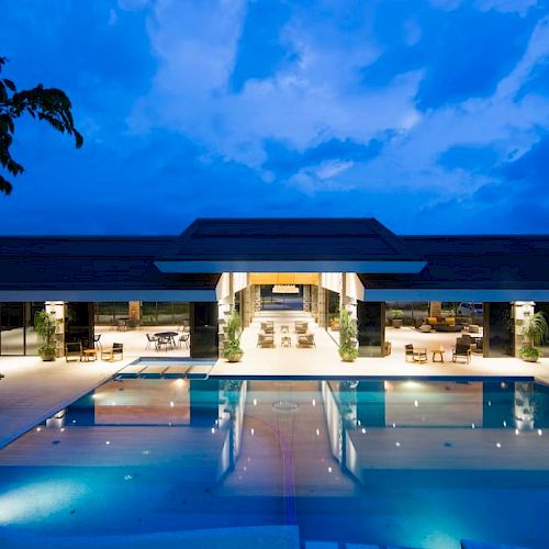A luxurious modern building features an illuminated outdoor swimming pool, surrounded by trees, with a clear evening sky in the background.