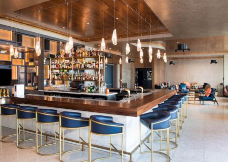 A modern bar with wooden accents, high shelves stocked with various bottles, blue bar stools, and unique pendant lighting, in a spacious lounge area.