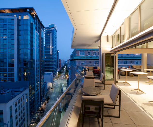 A modern balcony overlooking a city street with tall buildings. The balcony has several tables and chairs, and the view is at twilight.