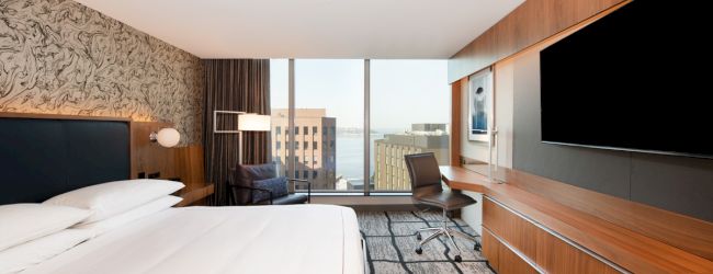 A modern hotel room features a large bed, flat-screen TV, desk, chair, and a window with a cityscape view in the background.