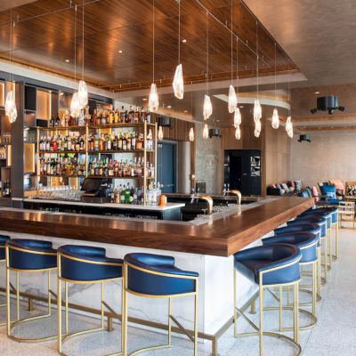 A modern, upscale bar with a sleek wooden counter, blue chairs, hanging lights, an extensive alcohol selection, and a cozy seating area in the background.