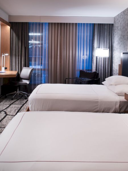 The image shows a modern hotel room with two neatly made beds, a mounted TV, a work desk with a chair, and contemporary decor with a stylish carpet and wallpaper.