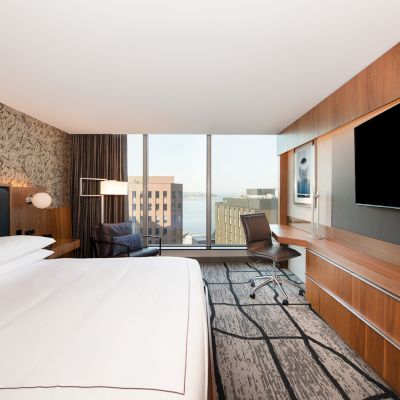 A modern hotel room featuring a large bed, a wall-mounted TV, a desk with a chair, and a cityscape view through large windows ends the sentence.