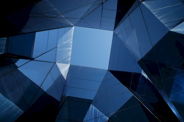 The image shows a view looking up at modern, reflective glass buildings forming a geometric pattern with a clear blue sky visible in the center.
