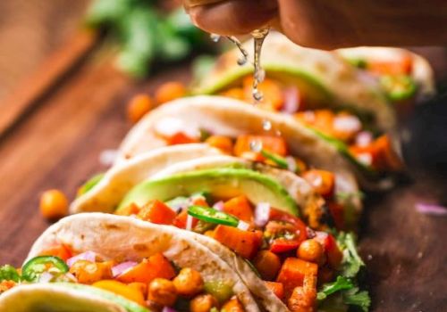 The image shows a hand squeezing lime over a row of colorful vegetable tacos on a wooden board, topped with chickpeas, avocado, and garnished with cilantro.