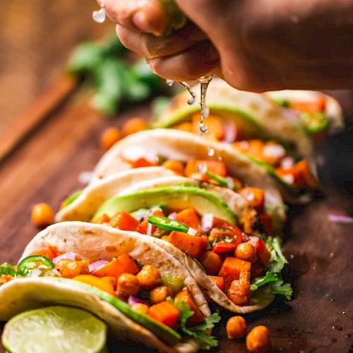 The image shows a hand squeezing lime over a row of colorful vegetable tacos on a wooden board, topped with chickpeas, avocado, and garnished with cilantro.