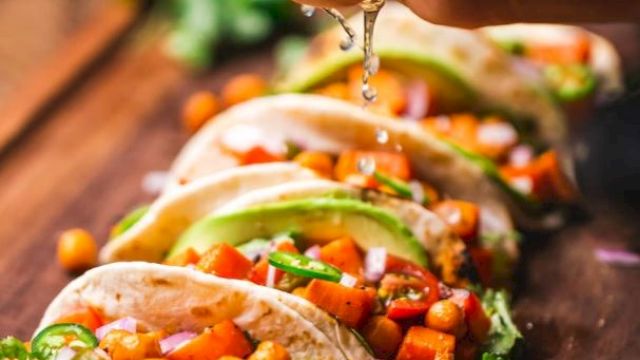 The image shows a hand squeezing lime over a row of colorful vegetable tacos on a wooden board, topped with chickpeas, avocado, and garnished with cilantro.