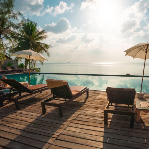 The image shows a serene beachside scene with lounge chairs, umbrellas, a wooden deck, a pool, and a view of the ocean under a partly cloudy sky.