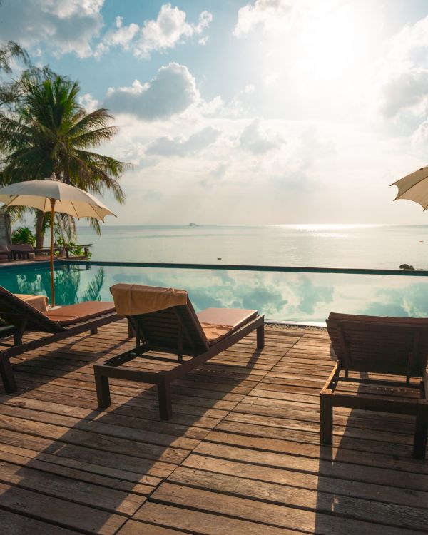 The image shows a serene beachside scene with lounge chairs, umbrellas, a wooden deck, a pool, and a view of the ocean under a partly cloudy sky.