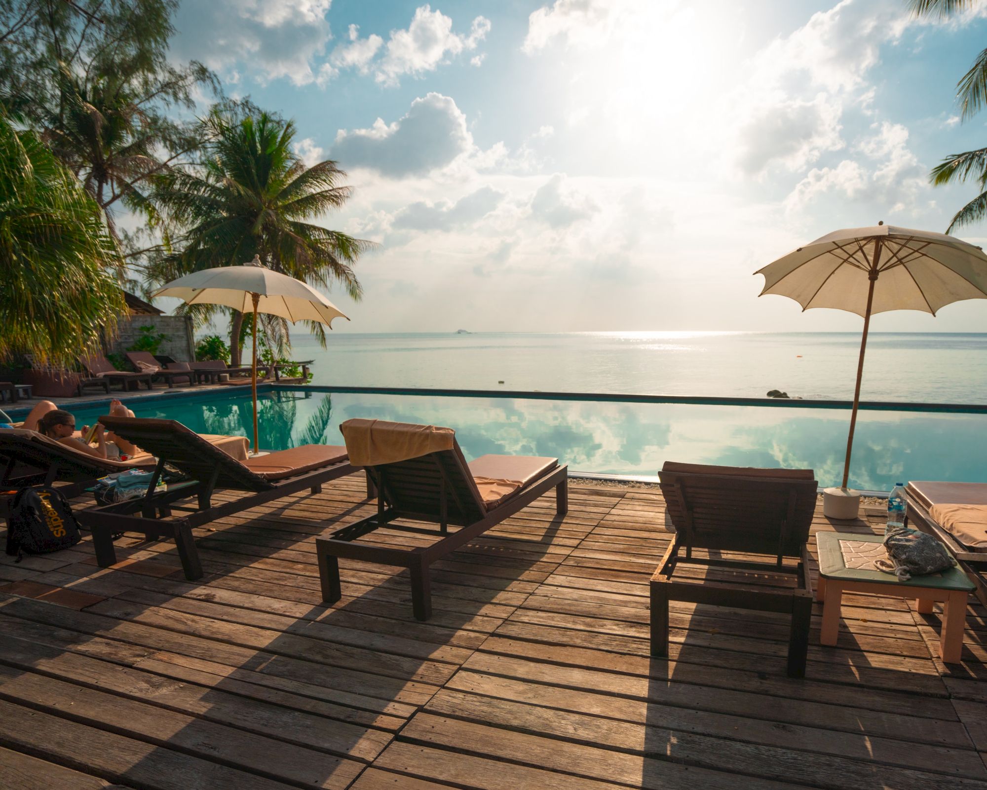 A serene poolside view overlooking the ocean with lounge chairs, umbrellas, palm trees, and calm waters reflecting the sky at dawn or dusk always.