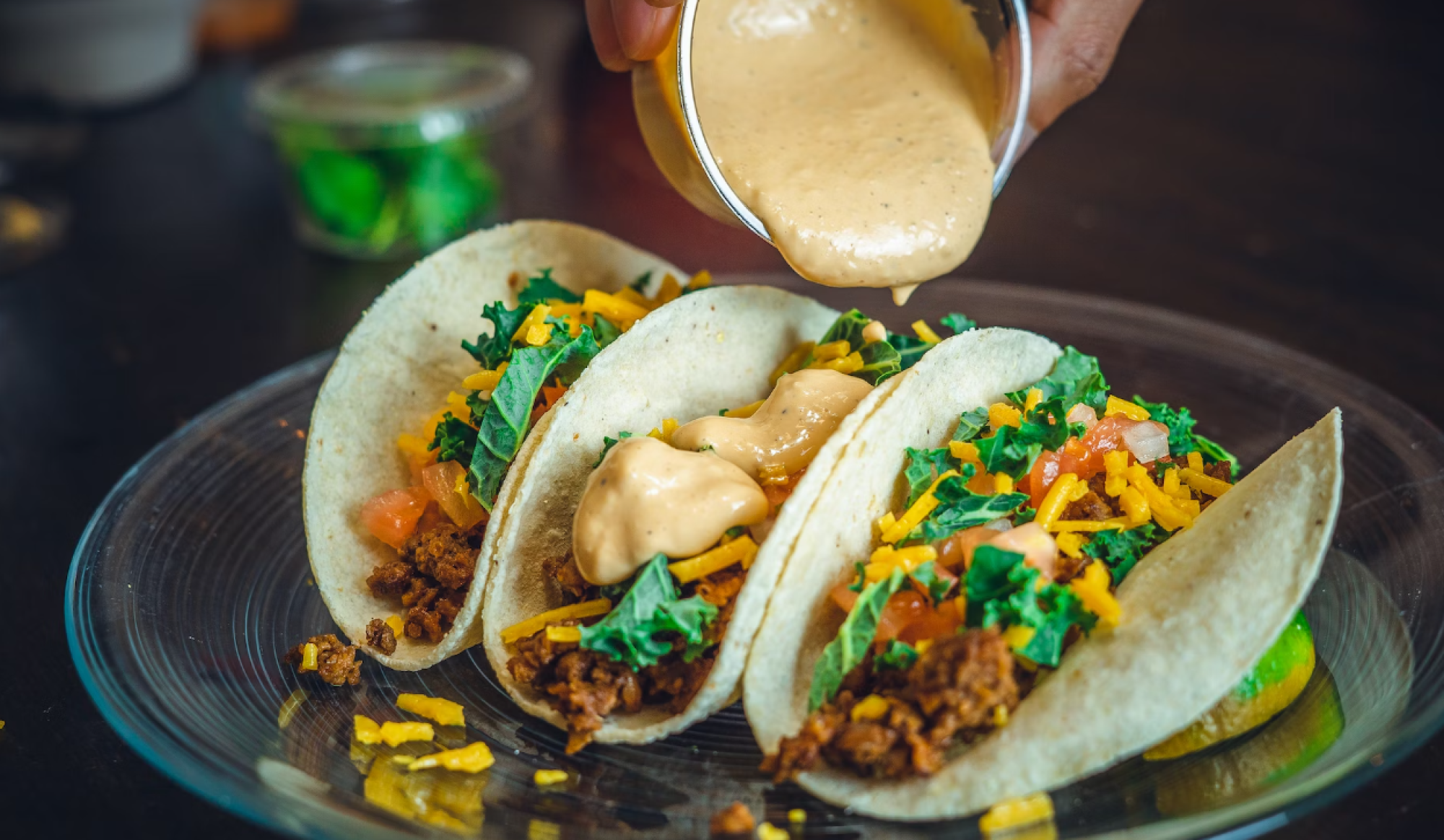 Three tacos loaded with ground meat, cheese, lettuce, and tomatoes, with a hand pouring sauce over them on a clear plate.