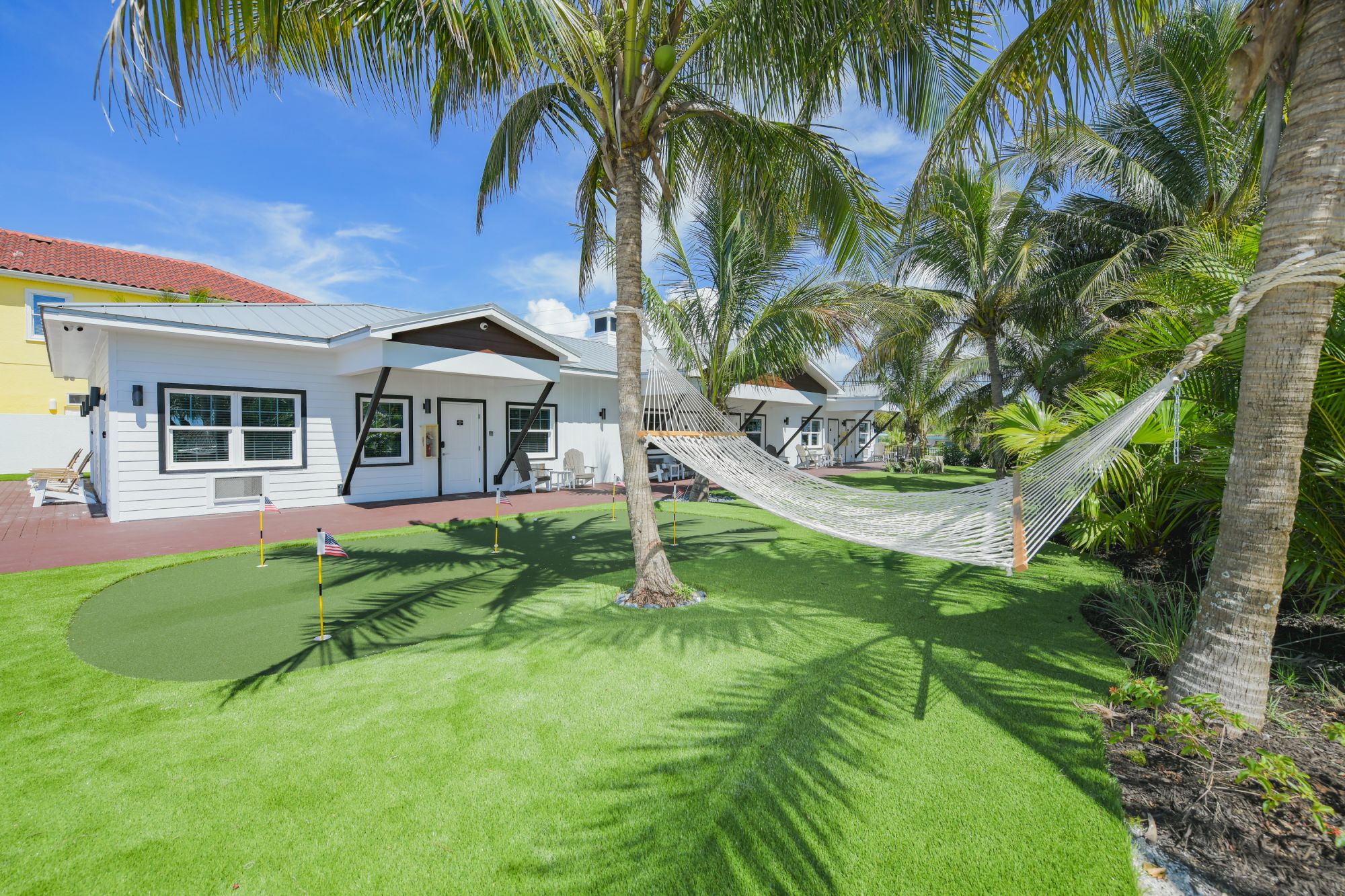 The image shows a relaxing scene with a hammock tied between two palm trees, a putting green, and a white house with a blue sky background.