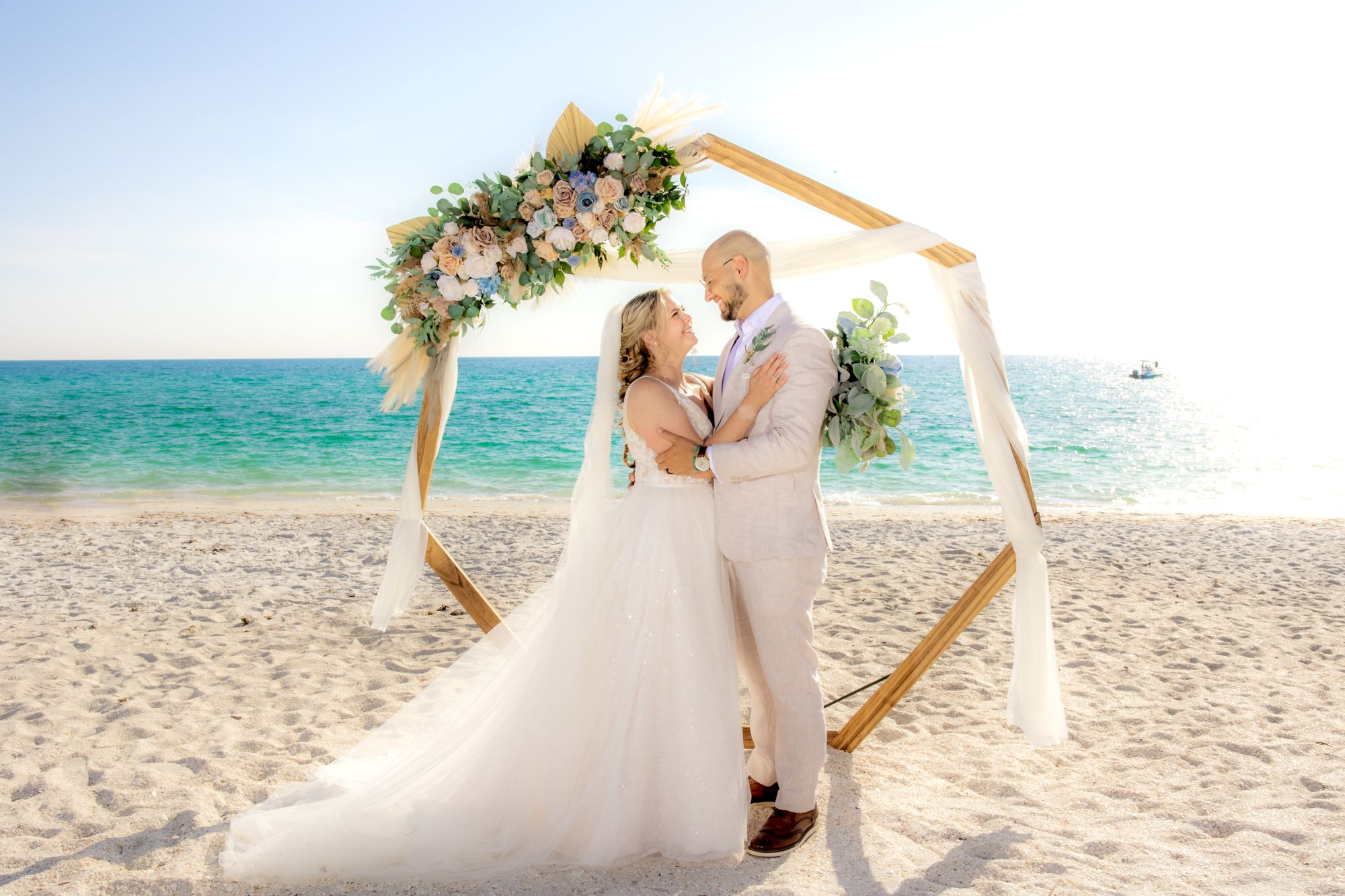 A bride and groom stand under a decorated hexagonal arch on a sandy beach with a turquoise ocean in the background, exchanging loving gazes.