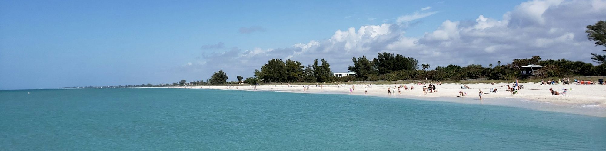 A serene beach scene with calm turquoise water, a sandy shoreline, a few people lounging, and clear blue sky with scattered clouds.