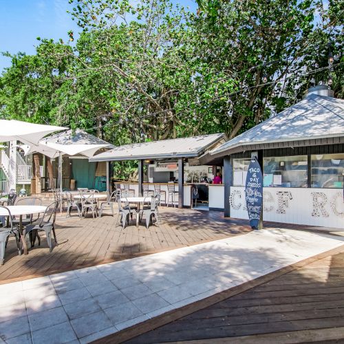 The image shows an outdoor café with tables, chairs, umbrellas, and a hut-style counter, surrounded by trees under a clear sky.