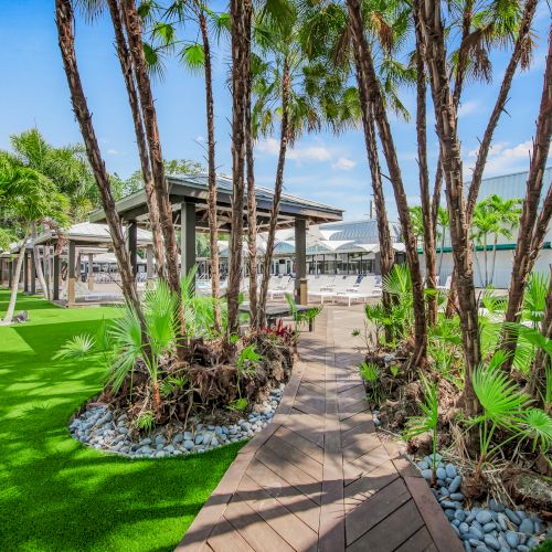 The image shows a tropical courtyard with palm trees, a wooden pathway, artificial grass, and nearby patios of white buildings in the background.