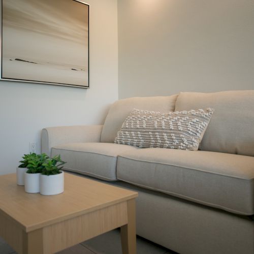 A modern living room features a beige couch, patterned pillow, wooden coffee table with plants, and a wall-mounted abstract painting.