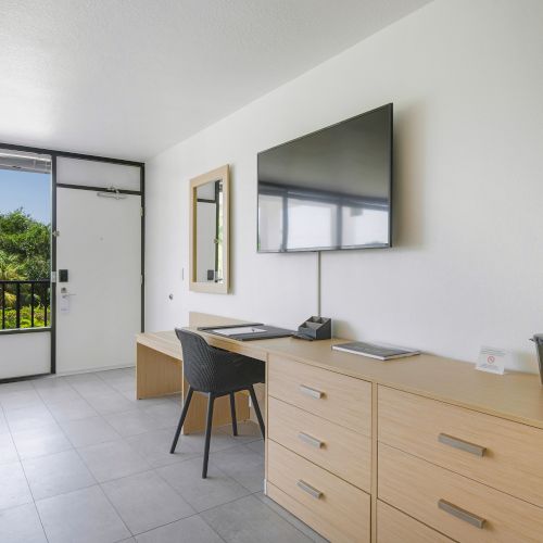 This image shows a minimalistic room with a desk and chair, flat-screen TV, large mirror, dresser, and a door leading to a balcony with greenery outside.