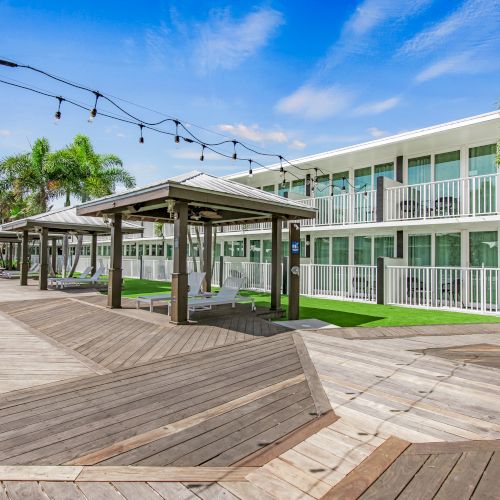 The image shows a modern, outdoor area with wooden walkways, pergolas, and string lights. A two-story building and palm trees are also visible.