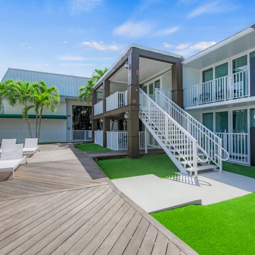 The image shows an outdoor courtyard with a wooden deck, lounge chairs, artificial turf, stairs, and a two-story building with balconies under a blue sky.