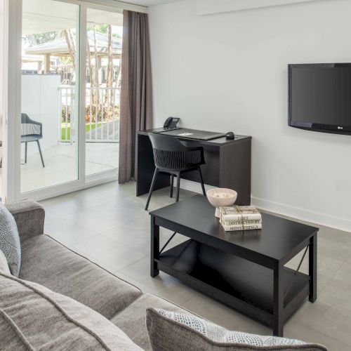 A modern living room features a sofa, coffee table, wall-mounted TV, desk with chair, and a sliding door opening to a balcony with outdoor seating.