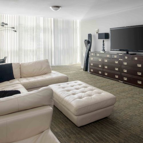 A modern living room with white sectional sofa, ottoman, large TV, lamps, and long dresser, featuring floor-to-ceiling blinds and desk.