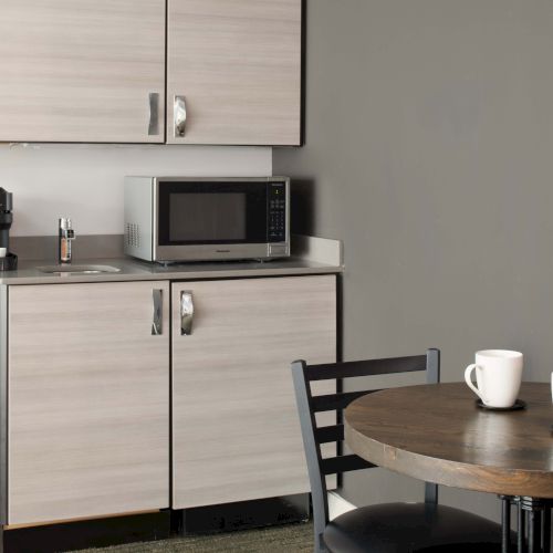 A small kitchenette with cabinets, a microwave, a coffee maker, and a round table with two chairs. Two white mugs are on the table.