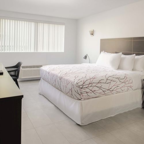 A neatly arranged bedroom with a bed, TV, chair, and nightstand beside a window with blinds, all in a minimalist style.