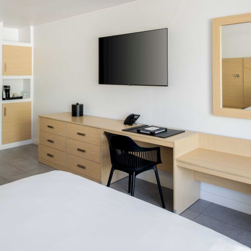 The image shows a modern hotel room with two beds, a flat-screen TV, a desk, a chair, a mirror, and a small kitchenette with a coffee maker.