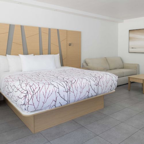 A modern hotel room with a large bed, abstract headboard, side tables, sofa, coffee table, wall art, tiled floor, and a partial view of the bathroom.