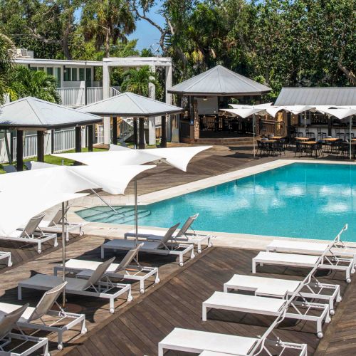 This image shows an outdoor pool area with white lounge chairs and umbrellas on a wooden deck, surrounded by greenery and shaded areas.