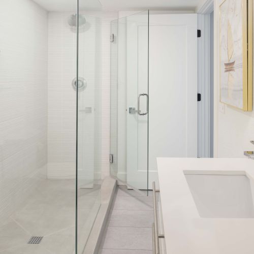 The image shows a modern bathroom with a glass-enclosed shower, a white countertop sink, a wall-mounted dispenser, a towel, and a framed picture.