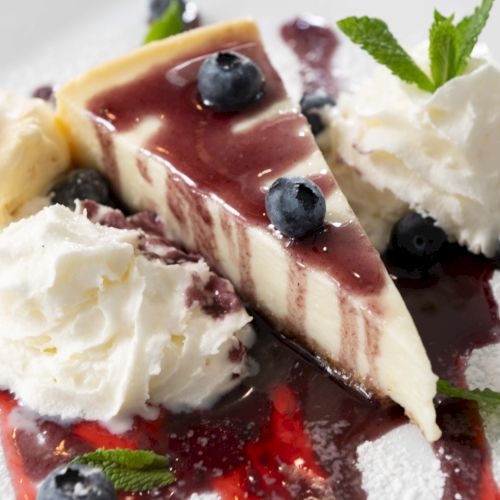 The image shows a slice of cheesecake with a berry sauce, whipped cream, blueberries, mint leaves, and a scoop of vanilla ice cream.