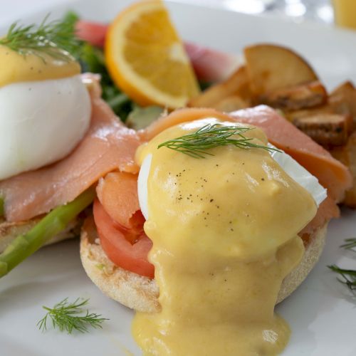 The image shows Eggs Benedict with hollandaise sauce, poached eggs, asparagus, ham on English muffins, and a side of potatoes and orange slices.