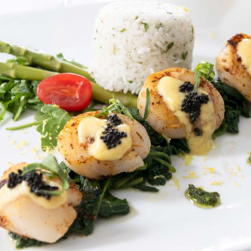 A plated dish featuring seared scallops topped with sauce and caviar, served with rice, asparagus, a cherry tomato, and leafy greens.