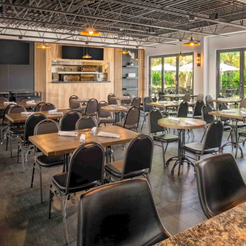 The image shows an empty, modern restaurant with neatly arranged tables and chairs, large windows, and an open kitchen area.