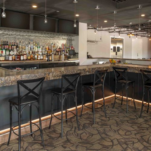 An empty bar features a granite countertop, high black chairs, wall hangings, and an array of bottles and glasses behind the bar.