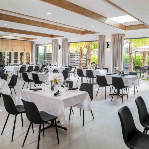 The image shows a well-lit dining area with multiple tables and chairs, set with white tablecloths and black seats, and large windows.