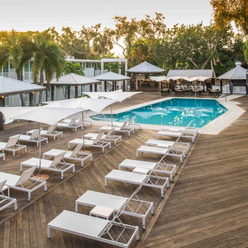 A serene poolside area with white lounge chairs, umbrellas, and a wooden deck, surrounded by lush greenery and buildings.