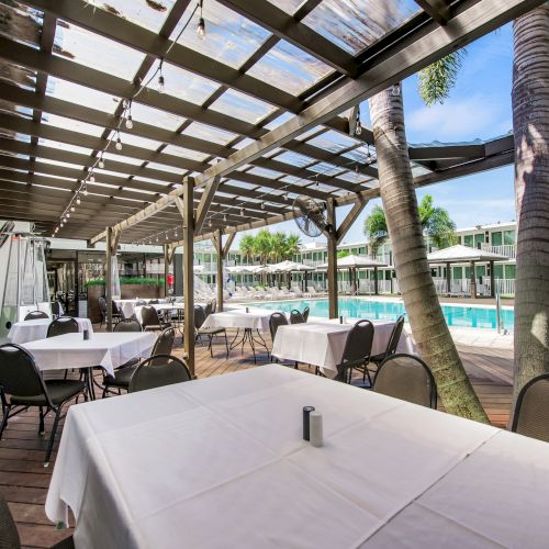 An outdoor dining area next to a swimming pool, under a pergola with tables and chairs, surrounded by palm trees and buildings in the background.