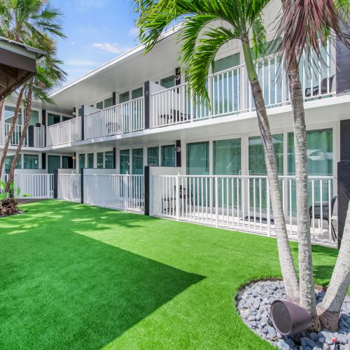 A modern apartment building with white railings, lush green courtyard, and palm trees under a clear blue sky is in the image.